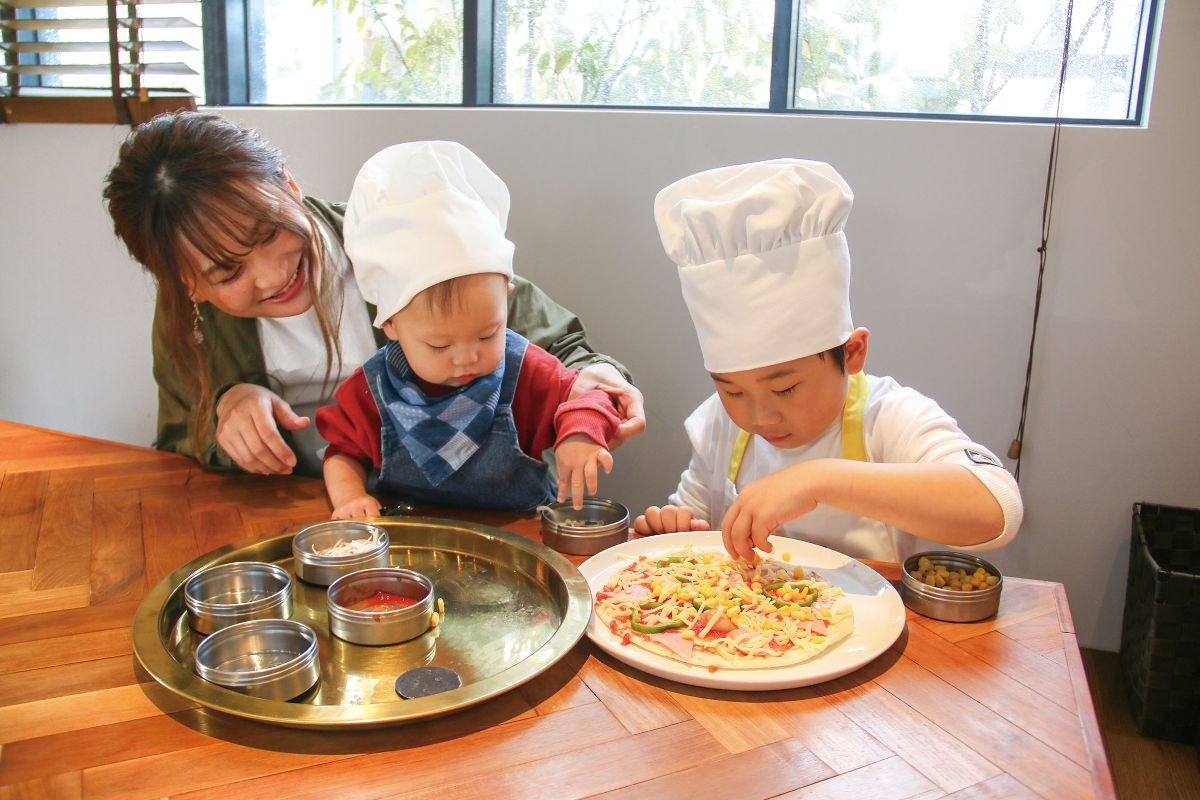 写真：テーブル席でキッズピザを作っている様子。右にピザクラストに具材を並べている4,5歳程度の男の子、左に1歳程度の赤ちゃんを抱いて男の子の手元を見守る母親。子供たちはコック帽をかぶっている