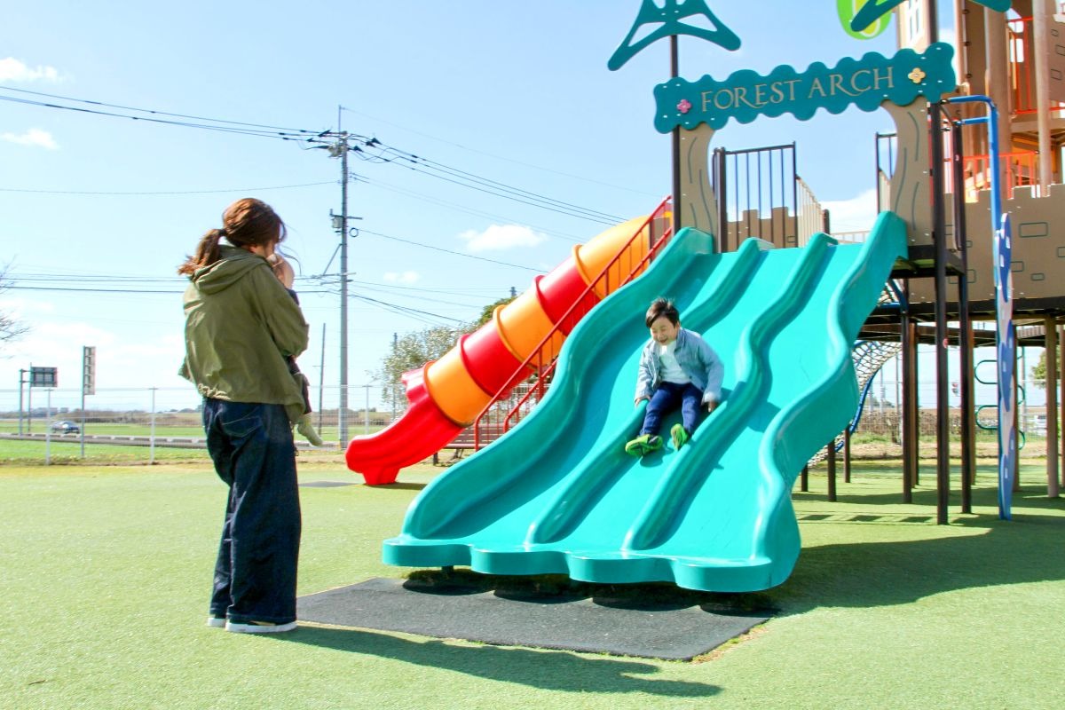 写真：大型遊具の滑り台を4,5歳程度の男の子が笑顔で滑り降りている。降り口で赤ちゃんを抱いた母親が見守っている