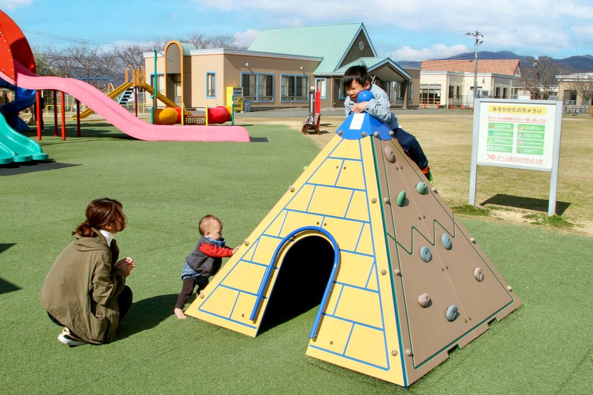 写真：手前の面に開口部があり、小さい子供が出入りできるピラミッド型の遊具。4，5歳程度の男の子が遊具の頂上付近に乗り、1歳程度の赤ちゃんが遊具のそばに立っている。母親が見守っている。