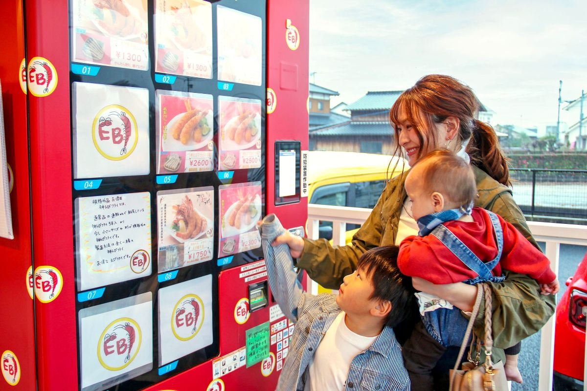 写真：自動販売機で冷凍えびフライを購入する親子の様子。4,5歳程度の男の子が自動販売機のボタンを押している。そばに立っている母親は左手で赤ちゃんを抱いて、右手を男の子のボタンを押す手に添えている