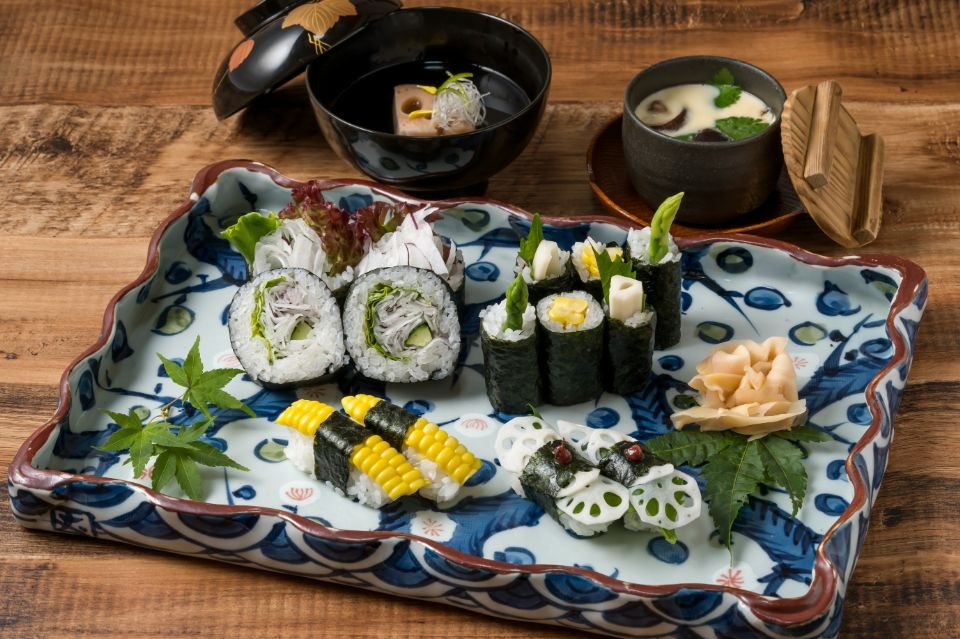 手前にトウモロコシ寿司、レンコン寿司、レタスの巻きずしが乗った四角の皿があり、奥に茶わん蒸し、お吸い物が並んだ料理画像