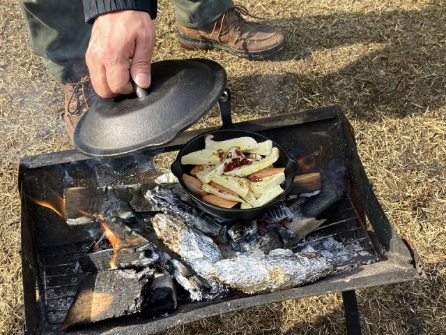 写真：焚き火台にセットしたスキレットの蓋を開けている男性の手元。スキレットの中にチーズ、ソーセージ、赤唐辛子が入っている