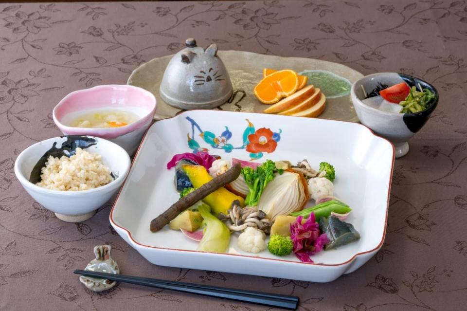 写真：手前に野菜料理が盛られた大きめの皿、左側にごはん、奥に輪切りの柑橘類と櫛切りのりんご盛り合わせと香の物がある様子