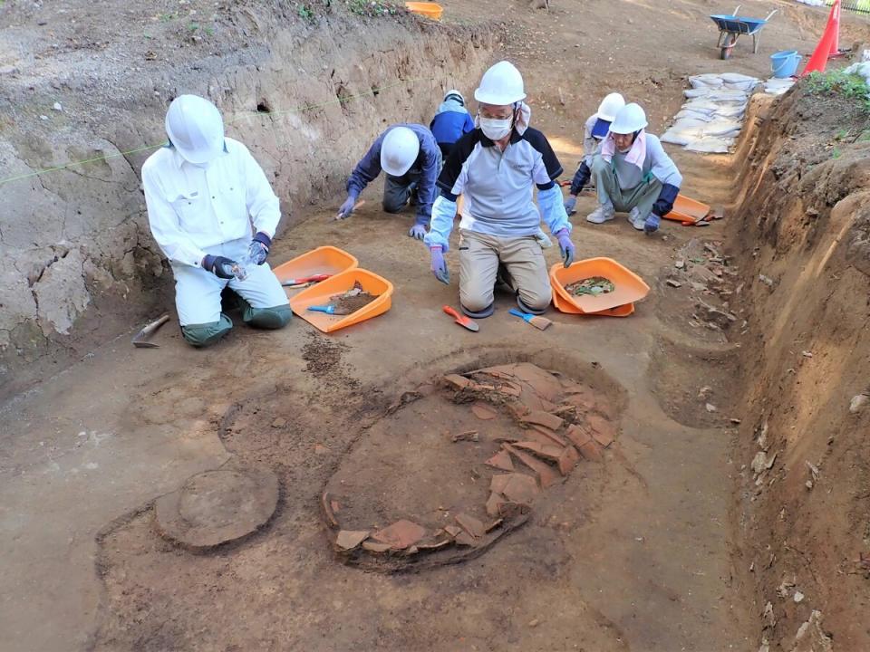 写真：甕棺が発掘されている現場。手前に甕棺の破片があり、発掘作業者２名が膝まづいて破片を見ている