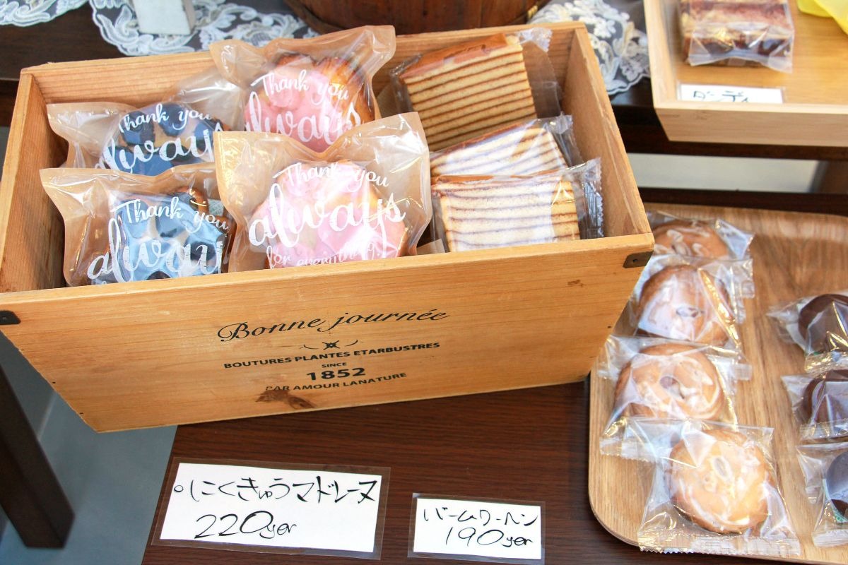 写真：焼き菓子が並んだ商品棚の様子。木箱に入ったマドレーヌ、バームクーヘンがある。その右側にトレイがありクッキーが並べられている