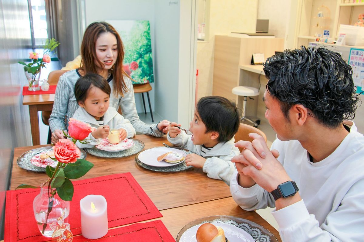 写真：テーブル席でお菓子を食べる家族。手前に父親、その隣に小学校低学年の子供。奥に母親と膝の上に3.4歳程度の子供が座っている