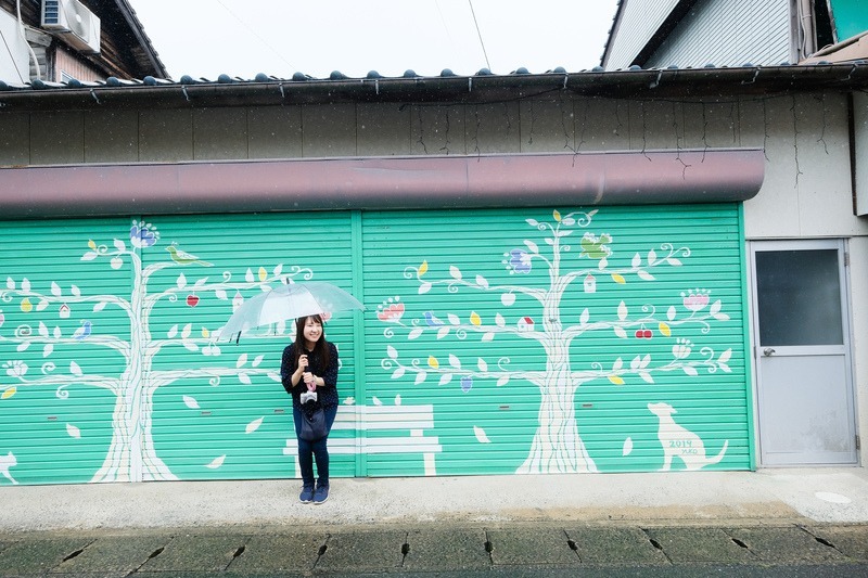 写真：木のイラストのウォールアートの前に女性が立っている写真