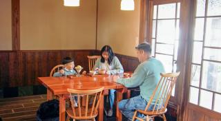 写真：テーブル席で食事する家族。手前に父親、奥に母親と３、４歳くらいの子供が並んで着席している
