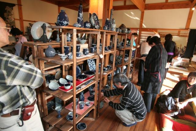 写真：木の棚に陶器が並べられ人々が陶器を手に取り見ている