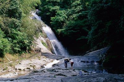 写真：水しぶきを上げる滝の遠景、手前の沢に人物が2人いる