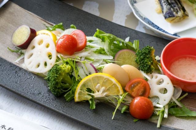 黒く四角の皿にレンコン、ブロッコリー、ミニトマトなどの野菜とドレッシングが添えられた画像