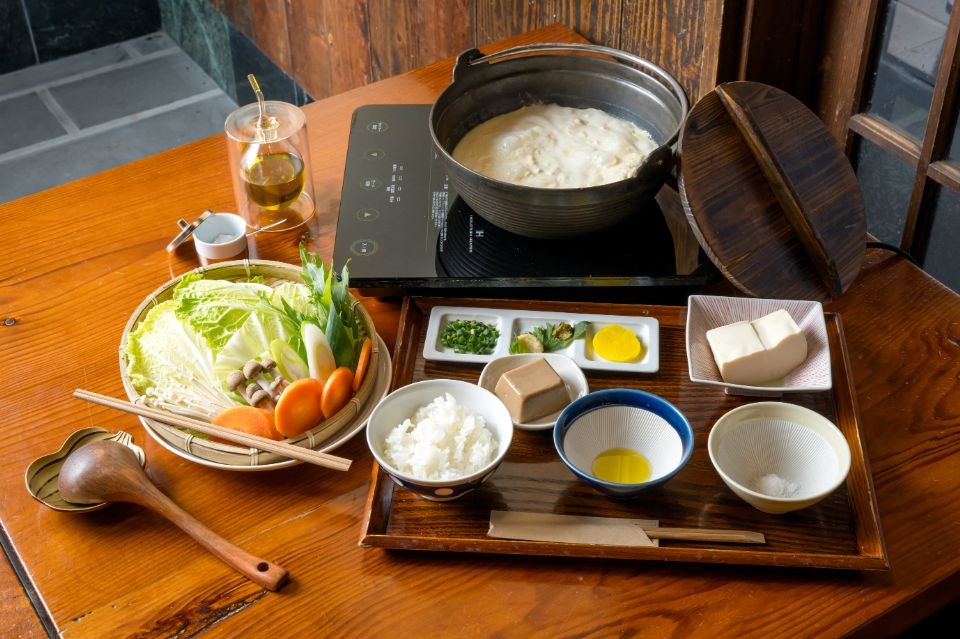 奥に鍋に入った温泉湯豆腐と手前に野菜盛合せ、塩、オリーブオイル、ご飯、豆腐、漬物が並べられた画像