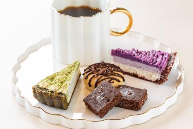 白い皿の上に緑色と紫色のケーキ、2種類のクッキー、カップに入ったコーヒーが乗ったデザート盛合せの画像