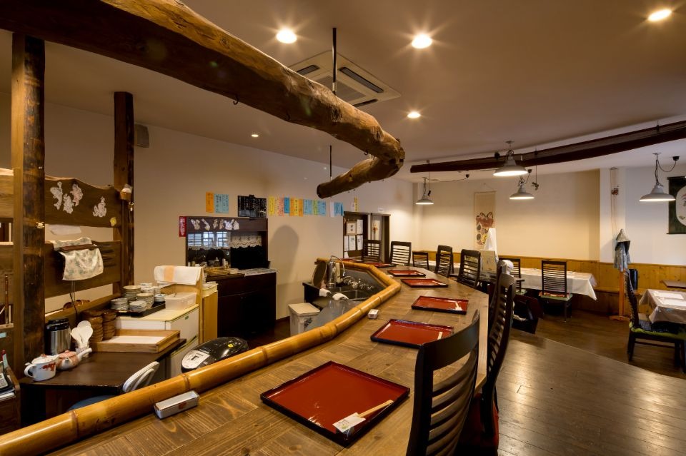 ベージュの壁と天井に、左側に木製で出来たカウンターと椅子が並ぶ店内画像