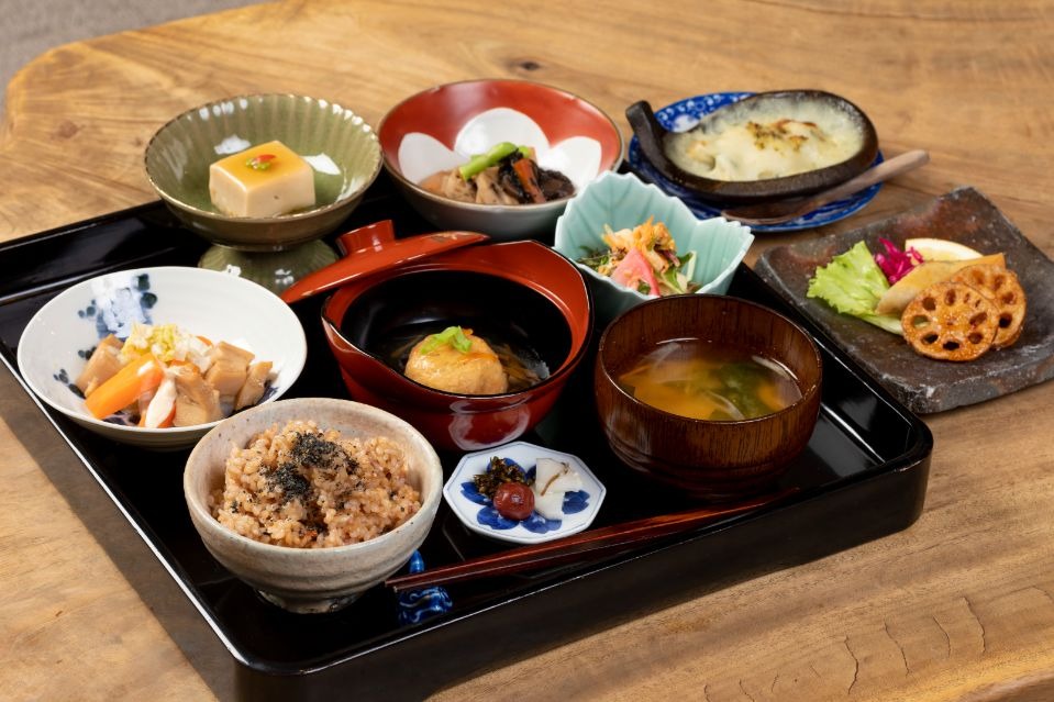 黒の盆の上に玄米ご飯、味噌汁、野菜を使った料理が複数並んだ料理の画像