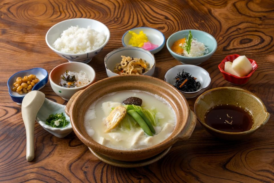手前に温泉湯豆腐、奥左から野菜の小鉢、ごはん漬物、調味料がテーブルにある画像