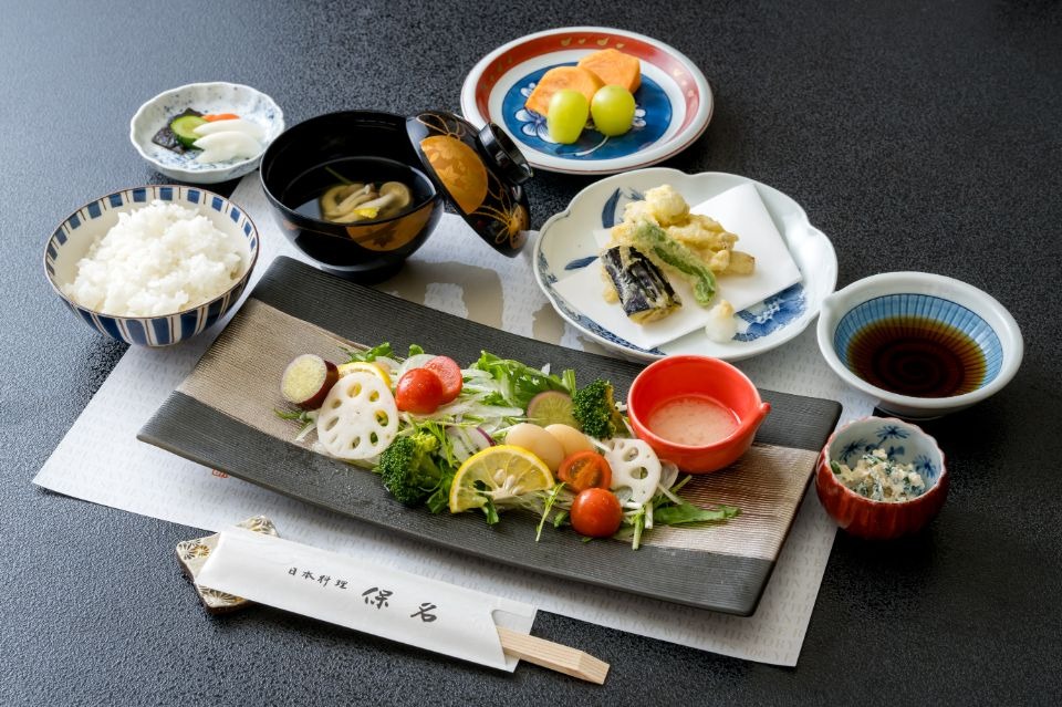 手前真ん中に野菜盛合せとドレッシングがあり、奥左からご飯、味噌汁、果物、天ぷら、つゆが配置された画像